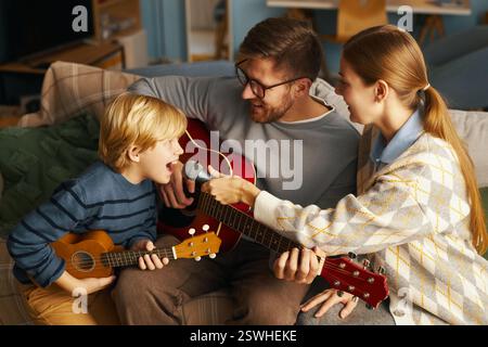 Die Familie sitzt auf der Couch, spielt Gitarren und singt fröhlich, Eltern teilen fröhliche Momente mit dem Kind in einem warmen und gemütlichen Wohnbereich Stockfoto
