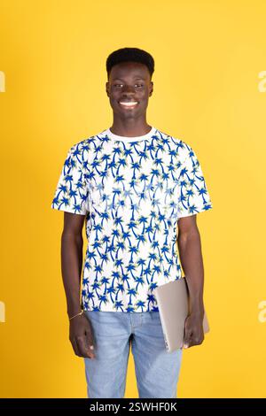 Ein junger Mann, der ein Hemd mit Palmen trägt, hält einen Laptop. Er lächelt und ist glücklich Stockfoto