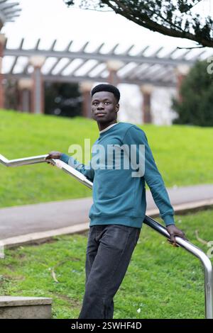 Ein junger Mann in einem blauen Pullover steht auf einem grasbewachsenen Hügel und lehnt sich auf ein Geländer Stockfoto