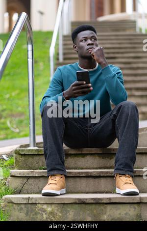 Ein junger Mann sitzt auf einer Treppe mit seinem Handy in der Hand. Er ist tief in Gedanken und denkt vielleicht über etwas Wichtiges nach Stockfoto