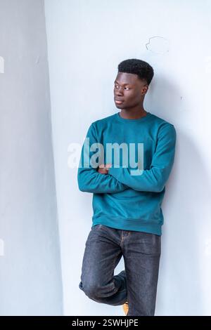 Ein junger Mann in blauem Sweatshirt und Jeans steht vor einer weißen Wand. Er blickt mit gekreuzten Armen in die Ferne. Das Konzept der Introspe Stockfoto