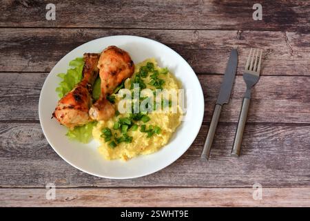 Ein Teller Kartoffelpüree mit grünen Zwiebeln und zwei gebackenen Hähnchenschenkeln auf einem Holztisch, neben einer Gabel und einem Messer. Draufsicht, flach. Stockfoto