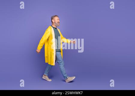 Lächelnder älterer Mann in gelbem Regenmantel, der auf lila Hintergrund geht und Freude und Stil ausstrahlt Stockfoto