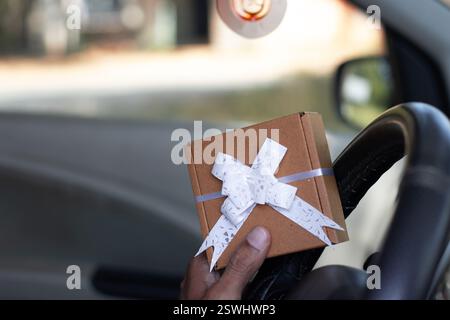 Der Mann hält eine braune Geschenkbox mit weißem Bogen auf einer Autosteuerung, valentinstag, frauentag, Muttertagskonzept Stockfoto