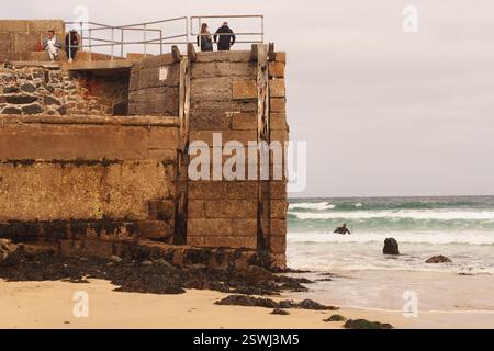 Urlauber stehen am Ende von New Pier St Ives, Cornwall, England, Großbritannien, mit Blick auf das Meer, die Wellen und ein Surfer auf dem Meer Stockfoto