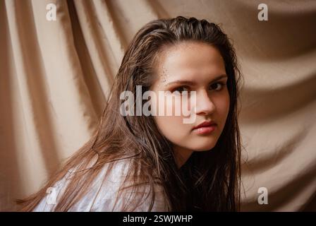 Intensives Porträt einer nachdenklichen jungen Frau vor einem drapierten Stoffhintergrund. Kaukasische Frau mit Glitzer im Gesicht. Beigefarbener Hintergrund. Stockfoto