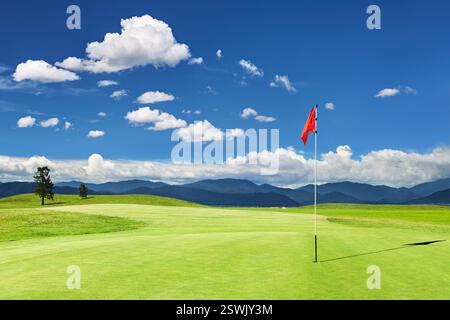 Schöner grüner Golfplatz mit Bergen, blauem Himmel und roter Flagge in einem Loch Stockfoto