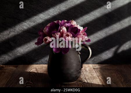 Das Foto zeigt einen Tulpenstrauß in einer dunklen Keramikvase auf einem Holztisch. Stockfoto