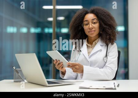 Porträt einer ernsthaften, selbstbewussten Ärztin am Arbeitsplatz in der modernen Klinikpraxis. Ärztin, die in die Kamera schaut, mit einem Tablet-Computer in den Händen, der am Schreibtisch sitzt. Stockfoto