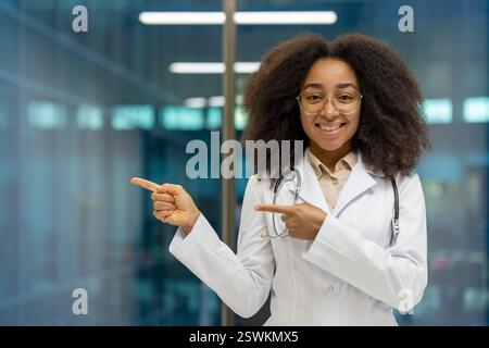 Eine freundliche afroamerikanische Ärztin in weißem Mantel und Stethoskop zeigt in einem modernen Krankenhaus auf die linke Seite des Rahmens Stockfoto