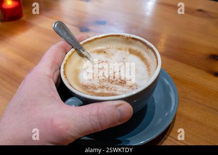 Eine Hand, die eine Kaffeetasse mit einem Löffel hält. Der Kaffee ist braun und mit Zimt bestreut Stockfoto