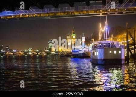 Nachtblick auf die Houses of Parliament und Big Ben, London, mit beleuchteten Flussbooten auf der Themse. Stockfoto