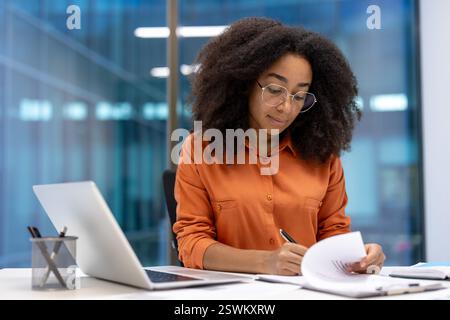 Ernsthaft fokussierte Frau, die Papierkram im Büro macht. Geschäftsfrau schreibt Daten auf, erstellt Finanzberichte. Buchhalter Finanzier sitzt am Schreibtisch mit Laptop. Stockfoto