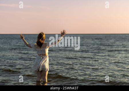 Eine Frau steht im flachen Wasser, die Arme vor Freude erhoben, während sie den ruhigen Sonnenuntergang bewundert. Der Horizont leuchtet in warmen Farben, die sich auf dem Meer spiegeln Stockfoto