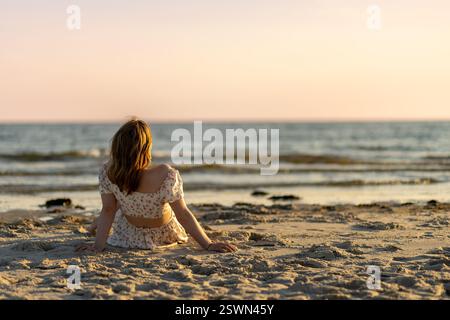 Eine Frau sitzt an einem Sandstrand, trägt ein leichtes Kleid und beobachtet den Sonnenuntergang über den ruhigen Meereswellen. Der Himmel leuchtet in warmen Farben, während sie mich reflektiert Stockfoto