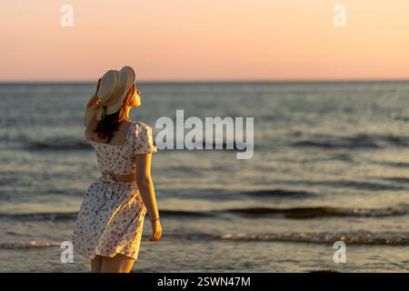 Eine junge Frau steht am Ufer, trägt ein weißes Blumenkleid und einen Strohhut. Sie blickt nachdenklich auf das Meer, während der lebhafte Sonnenuntergang das zeigt Stockfoto