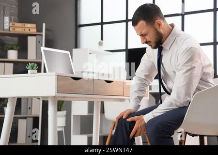 Junger Geschäftsmann mit Knieschmerzen am Tisch im Büro Stockfoto