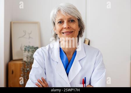 Porträt einer reiferen Ärztin mit Blick auf die Kamera, die im Büro auf dem Schreibtisch sitzt Stockfoto