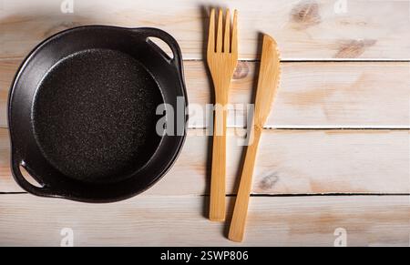 Leere schwarze Keramikplatte im Pfannenstil neben Holzbesteck auf einem rustikalen weißen Holztisch, Blick von oben. Stockfoto