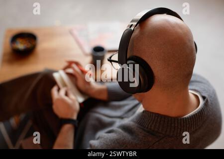 Rückansicht eines Glatzköpfigen Mannes, der Musik mit Kopfhörern hört, um die Konzentration beim Schreiben im Notebook zu verbessern, am Couchtisch im Heimbüro, im Kopierraum Stockfoto