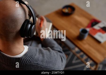 Rückansicht eines Mannes mit rasiertem Kopf, der Musik mit intelligenten kabellosen Kopfhörern hört, während er in einem Notebook am Couchtisch im modernen Büro schreibt, Kopierraum Stockfoto
