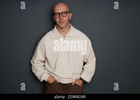 Mittelgroße Aufnahme eines selbstbewussten Mannes mittleren Alters mit rasiertem Kopf und weißem Sweatshirt mit halbem Reißverschluss, das modisches Outfit im Studio und im Kopierraum an der grauen Wand zeigt Stockfoto