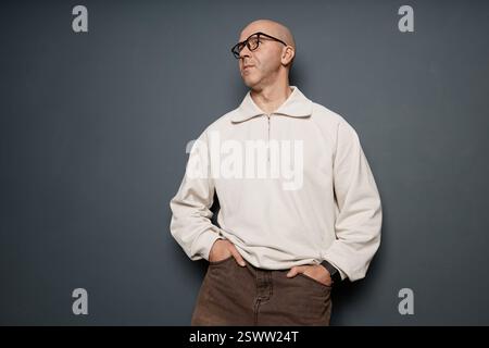 Mittelgroße Aufnahme eines Mannes mittleren Alters mit Buzzcut in weißem Sweatshirt mit halbem Reißverschluss, der mit Händen in den Taschen gegen die graue Wand im Studio posiert, im Kopierraum Stockfoto