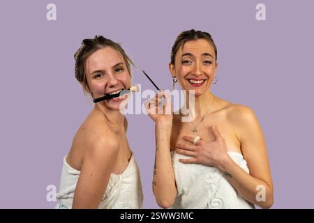 Zwei fröhliche junge Frauen, die in Handtücher gewickelt sind, genießen einen Moment der Schönheit und des Lachens und halten Make-up-Pinsel vor einem lila Hintergrund Stockfoto