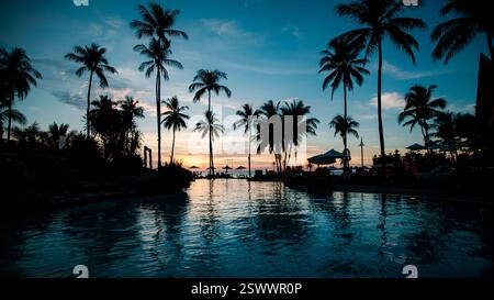 Ein tropischer Strand mit Palmensilhouetten, die sich im Wasser spiegeln und unter einem atemberaubenden Sonnenuntergang glühen. Stockfoto