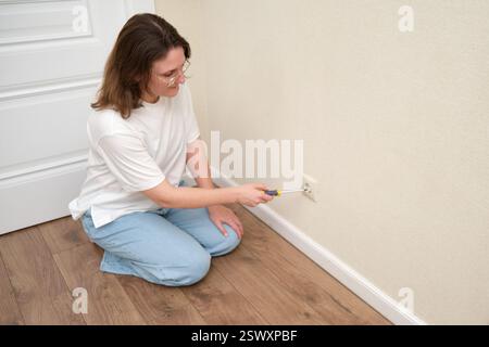 Frau repariert die Steckdose mit einem Schraubenzieher an der beigefarbenen Wand. Hauswartung und elektrisches Arbeitskonzept Stockfoto