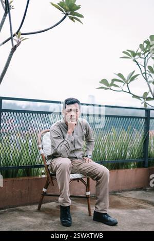 Ein nachdenklicher Mann sitzt auf einem Stuhl und betrachtet das Stadtbild. Er wirkt nachdenklich in seiner Haltung und findet einen Moment des Friedens in der Mitte Stockfoto