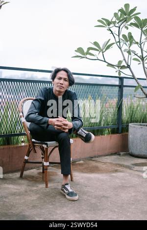Ein nachdenklicher junger Mann sitzt draußen auf einem Stuhl und sieht ruhig und stilvoll aus. Er trägt lässige Kleidung, mit gekreuzten Armen, und sein Ausdruck zeigt sich Stockfoto