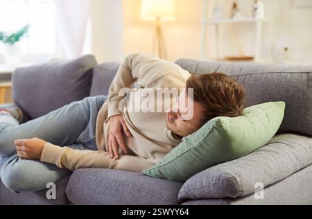 Junger kranker Mann, der seinen Bauch berührt und an Magenschmerzen leidet, der zu Hause auf dem Sofa liegt. Stockfoto