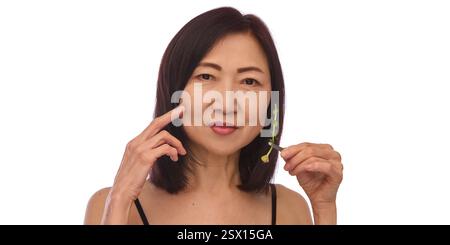 Schöne koreanische Frau in ihren 60ern, die mit einer Hand das Gesicht berührt, grüne Pflanze mit der anderen hält und ein schwarzes Tanktop vor weißem Studiohintergrund trägt. Stockfoto