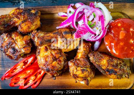 Saftige gegrillte Hähnchenkeulen werden auf einer Holzplatte serviert, begleitet von leuchtenden roten Zwiebeln, frischen Chili-Scheiben und einer würzigen Sauce. Ein Delicio Stockfoto