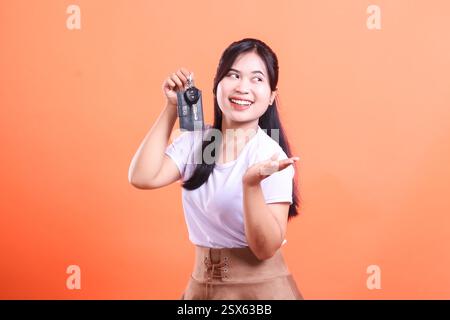 Aufgeregte asiatische Frau, die ihre neuen Autoschlüssel mit einem großen Lächeln zeigt. Zur Seite schauen und präsentieren. Isoliert auf orangefarbenem Hintergrund Stockfoto