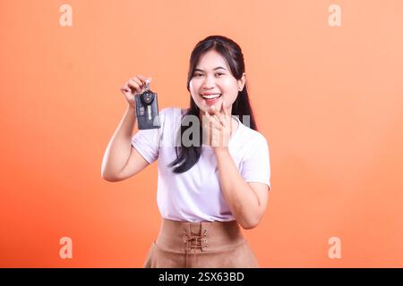 Eine lächelnde junge asiatische Frau, die einen Autoschlüssel in der Hand hält. Sie sieht glücklich und selbstbewusst aus mit ihrem neuen Fahrzeug auf orangefarbenem Hintergrund Stockfoto