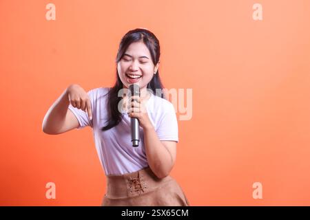 Eine junge Frau lächelt breit, während sie in ein Mikrofon singt und ihren Finger zur Seite zeigt, isoliert auf orangefarbenem Hintergrund Stockfoto