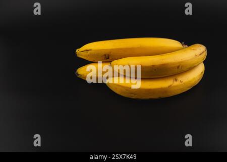 Vier reife Bananen sind übereinander gestapelt und zeigen ihre leuchtende gelbe Farbe. Der dunkle Hintergrund hebt ihre natürlichen Texturen hervor und c Stockfoto