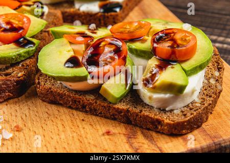 Farbenfrohe Sandwiches mit offenem Gesicht, mit frischer Avocado, Kirschtomaten und einem Hauch Balsamico-Glasur stehen auf einem hölzernen Servierbrett zur Schau Stockfoto