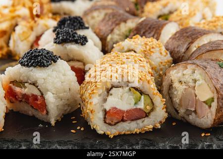 Eine Vielzahl von Sushi-Brötchen sind kunstvoll auf einem dunklen Steinteller angeordnet. Die Brötchen enthalten farbenfrohe Zutaten wie Fisch, Gemüse und Sesam Stockfoto
