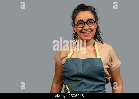 Porträt einer fröhlichen, reiferen Chefkochin mit Schürze und Brille, lächelnd auf grauem Hintergrund Stockfoto