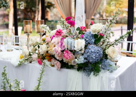 Ein atemberaubendes buntes Blütenpracht schmückt einen wunderschön gedeckten Tisch im Freien. Die Blumenarrangements sind mit Rosen, Dahlien und Hortensien gestaltet Stockfoto