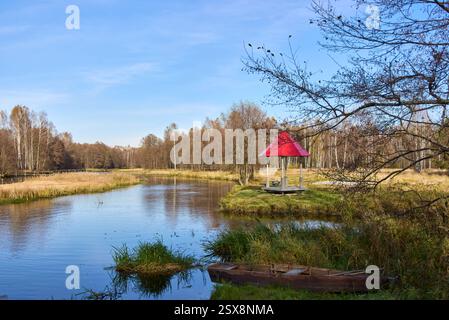 Eine ruhige Seeszene mit einem Pavillon mit rotem Dach, umgeben von Herbstbäumen und einem Holzboot am grasbewachsenen Ufer Stockfoto