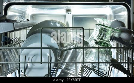 Detail des oberen Geschirrspülmaschinenregals mit Metallplatten, gefüllt mit frisch gewaschenen Tellern, Tassen und Gläsern mit Hintergrundbeleuchtung. Vorderansicht. Stockfoto