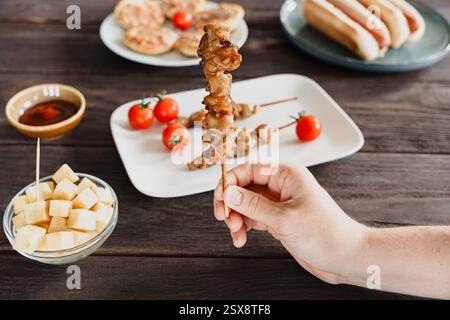 Eine Hand hält einen gegrillten Hähnchenspieß mit Barvorspeisen auf einem Holztisch Stockfoto