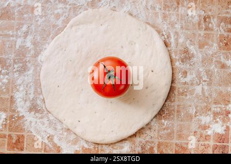 Ein ausgerollter Pizzateig mit einer frischen roten Tomate in der Mitte, hausgemachte Pizzazubereitung Stockfoto