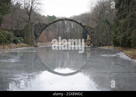 Kromlau, Deutschland. Februar 2025. Die Rakotzbrücke spiegelt sich in einem eisbedeckten See im Azalea- und Rhododendron-Park wider. Quelle: Sebastian Kahnert/dpa/Alamy Live News Stockfoto