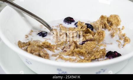 Eine Schüssel mit knusprigem Müsli und Heidelbeeren, die in Milch schwimmen, steht auf einem Esstisch. Ein silberner Löffel liegt drinnen, bereit für ein köstliches Frühstück Stockfoto
