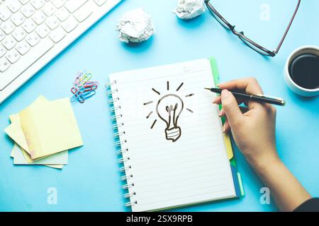 Frau zeichnet Leuchtbirne auf Notebook mit Tastatur und stationär auf Tisch Stockfoto